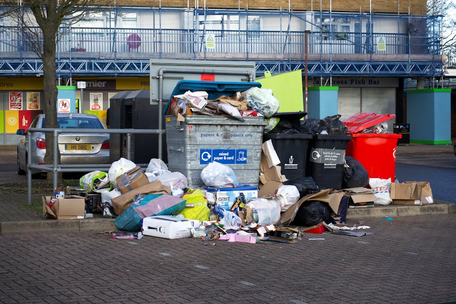 An overflowing collection of mixed waste and rubbish bags is piled up in front of a designated recycling and waste collection point on a paved urban street. The waste includes crumpled cardboard boxes